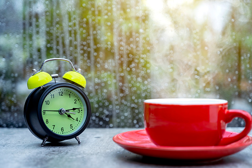 A coffee cup and alarm clock on the table with a clear window and raindrop background