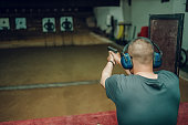 A professional man wearing earmuffs and goggles is practicing shooting a 9mm pistol inside a shooting range.