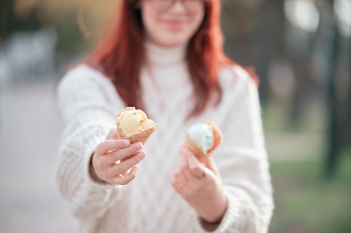 Girl in a white sweater holds out two ice cream cones.