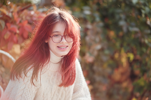 Beautiful long-haired teenage girl in glasses sits on a background of autumn foliage. Backlight, sunlight.