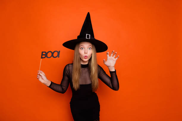 ritratto fotografico di una bella ragazza adolescente bionda che tiene il segno del boo graffia vestito a mano vestito nero di halloween isolato su sfondo di colore arancione - witch smiling evil bizarre foto e immagini stock