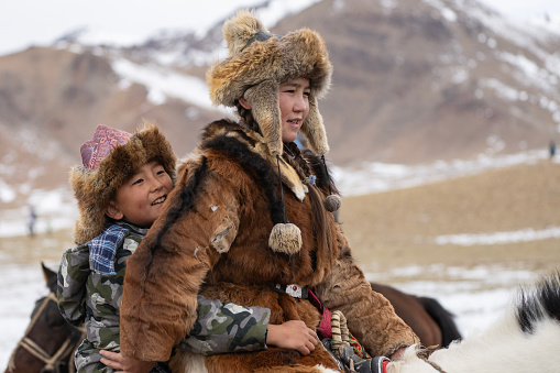 Bayan-Olgii Province, Mongolia - October 1, 2023: A boy hugs teenager Aimuldir D., an ethnic Kazakh female eagle hunter and eventual winner of the Ulgii Golden Eagle Festival, as they ride a horse.