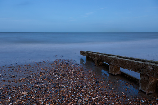 Wooden groyns in pebbly beach on the coast of Eastbourne. Calm and flat sea in the background.