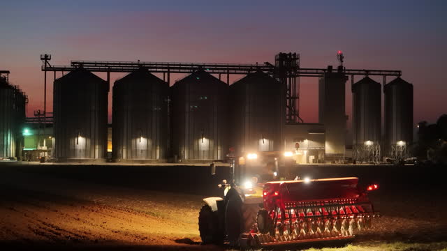 Illuminated Tractor Plowing Field with Corn Silos in Background at Night