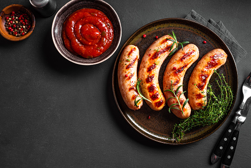 Grilled sausages with tomato sauce and herbs on black background, top view, copy space. Roasted homemade wurst sausages on plate.