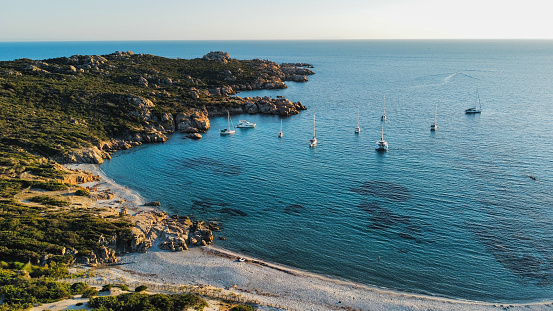 Aerial shot of a heavenly Corsican cove at sunset, with sailboats at anchor, rocks , turquoise water and a large beach.