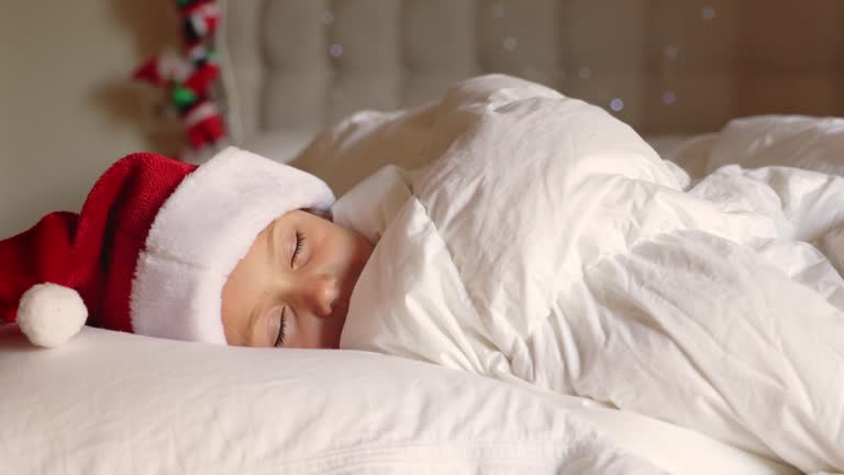 Little child girl wearing red Christmas pajamas and white fluffy Santa hat lie pillow under cozy warm blanket. Sleep at night and smile sweet dreams. Merry Christmas Happy New Year Holiday 2023