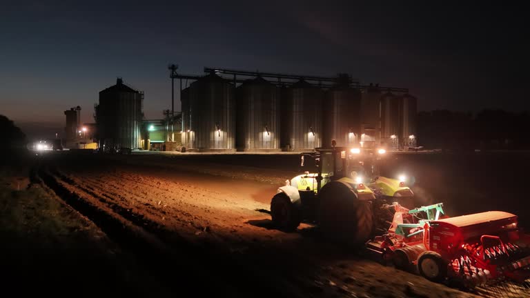 Farmworker Plowing Corn Field with Illuminated Silos in Background at Night