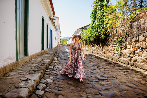 Smiling young woman wearing a dress and sun hat walking along a cobblestone street in a historic town center