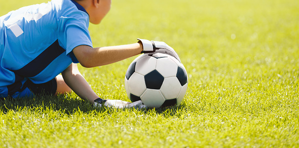 A young boy as a soccer goalie catching a soccer ball during a tournament game. A school football goalkeeper in a blue jersey shirt and sports gloves throwing the ball and playing a football match