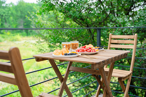 Lovely breakfast outdoors. Table set on the summer terrace. Fruits and berries, croissants and coffee on the table overlooking the green landscape.