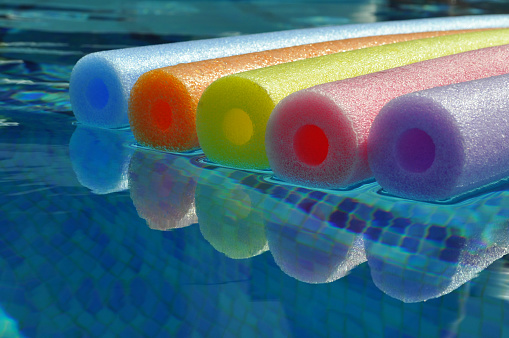 Rainbow coloured pool noodles floating in swimming pool, with reflection in water