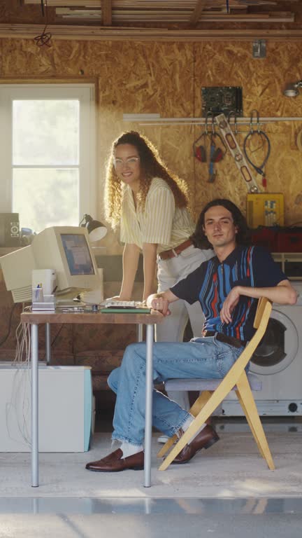 Vertical Screen: Caucasian Male Software Developer And Hispanic Female Designer Looking At Camera And Smiling In Retro Garage With Old Computer. Startup Founders Working On Online Service In Nineties.