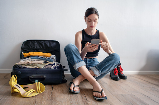 Happy young woman in colorful summer outfit sitting on staffed suitcase with smart phone