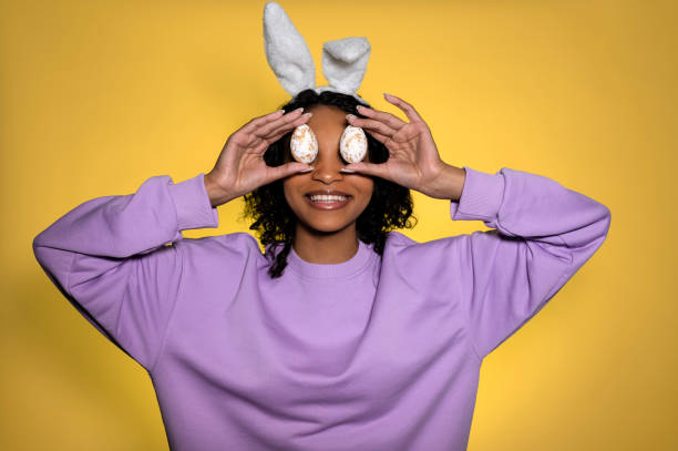 schöne dunkelhäutige frau, die mit hasenohren lächelt und bunte ostereier vor den augen hält - easter easter bunny fun humor stock-fotos und bilder