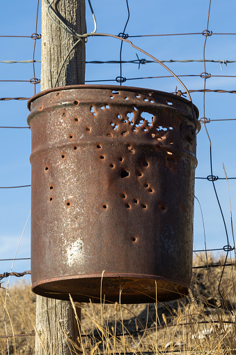 Close-up of a rusty old metal five gallon pail that has been used a target shooting practice is hanging from a wooden fencepost of a barbed wire fence.