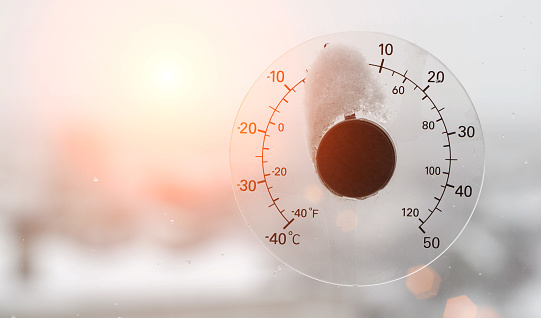 The rays of the setting sun in the sky against the background of a street thermometer with a snow cap on the window. Snowy weather and warming concept. Weather forecast