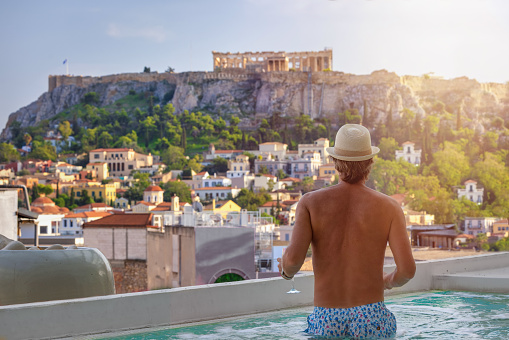 A tourist man in a swimming pool enjoys the view over the old town of Athens