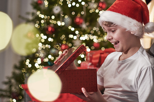 A joyful boy in a Santa hat opens a Christmas present. Christmas Eve Magic night