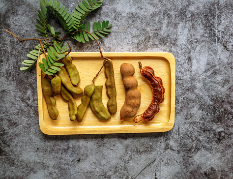 Close-up of ripe brown tamarind fruit and raw green tamarind pod with green leaf tamarind on a wooden tray gray background, brown fruit peel.