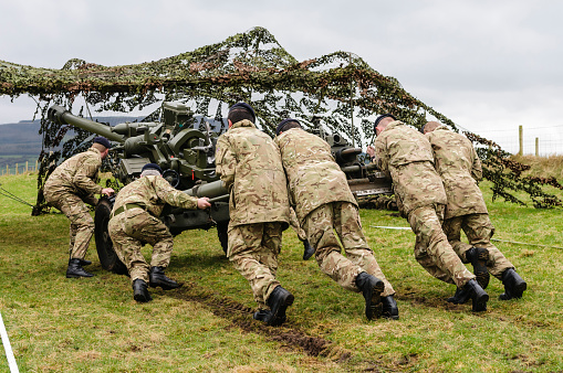 Magilligan Army Training Grounds, Londonderry, Northern Ireland, UK. 05/04/2014 -  A 6-man squad of soldiers from the Royal Artillery Corp of the British Army, wearing camouflage fatigues, regimental berets, and black boots push a 105mm Light Field Gun Howitzer into position underneath camouflage netting during a training exercise to hone their skills and increase speed of artillery deployment.  The soldiers were tasked with driving the gun to its intended location, unhitching it, transforming it to its firing configuration, positioning it below camouflage netting, aiming it using a grid position provided by a team of forward observers, and firing it before reversing the procedure and driving away.  All attempts by all squads were accurate to within 5m at 1.5km, and the fastest time to deploy, fire and return was 4m30s.