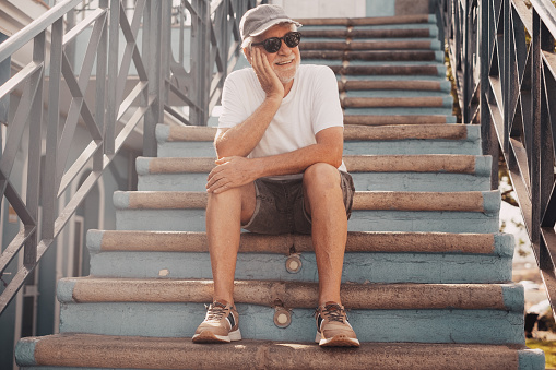 Portrait of senior handsome man smiling relaxed sitting in outdoors on a staircase. Carefree elderly man with hat and sunglasses enjoying vacation or retirement
