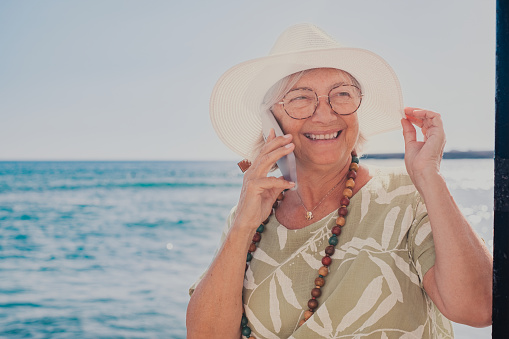 Portrait of handsome cheerful senior woman in white hat and eyeglasses talking using mobile phone sitting at the beach enjoying free time and vacation