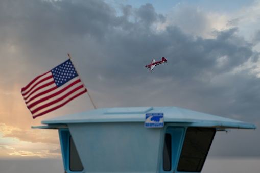 An airplane soars in the sky above a lifeguard station, with an American Flag fluttering in the wind in the foreground