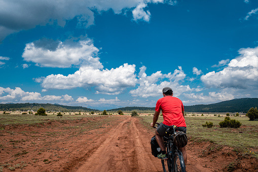 Kırmızı tişörtlü bisikletli.Bikepacking sistem.Doğada toprak yolda gidiyor.Arka plandan çekim.Model 31 yaşında Hava açık bulutlu.Doğa manzarası macera ruhu yüksek.