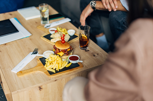 Tasty hamburger and French fries on a wooden board on a table in a restaurant, where two people are sitting