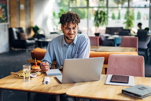 Male entrepreneur is on his lunch break using a laptop and writing notes in a notebook