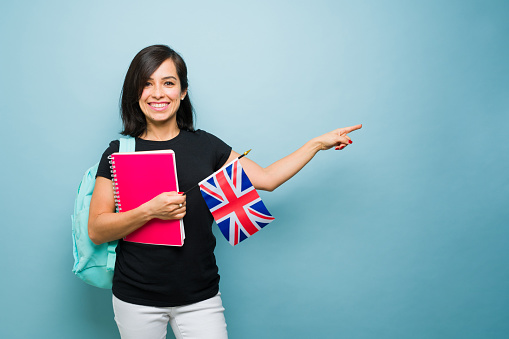 Beautiful woman pointing to copy space ad against a blue background while learning English with books holding a UK flag