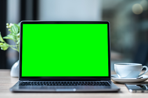 Mockup of laptop computer with empty screen with coffee cup and smartphone on table of the coffee shop background,Green screen