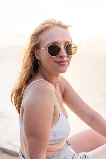 A striking stock image featuring a beautiful redhead in her 20s, set against the backdrop of a picturesque sunset by the water. The subject exudes a captivating charm while wearing stylish sunglasses, perfectly complementing the warm, golden hues of the evening.

In this photograph, the young woman's radiant beauty shines through as she takes in the serene atmosphere of the waterfront. Her sunglasses add a touch of contemporary flair to her look, and her natural grace harmonizes with the tranquil setting.

This image beautifully encapsulates the allure of youthful beauty and the serene ambiance of a waterfront sunset. It's an ideal asset for a wide range of creative projects that seek to capture the essence of a vibrant, redheaded woman at the magic hour by the water.