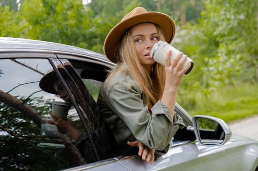 Smiling young woman looking from car window and drinking coffee or tea from reusable thermos cup. Local solo travel on weekends concept. Exited woman explore freedom outdoors in forest. Unity with nature lifestyle, rest recharge relaxation