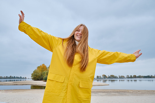 A happy girl in a yellow raincoat walks along the beach of a lake or river on a cool day. Travel concept. Girl on a journey. Amazing scenic street view