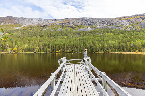 Woman enjoying peaceful environment in Norway. Lake and green mountains.\nShe relaxes on a wooden pier.