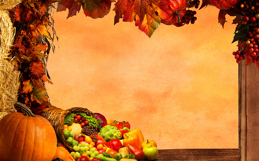 A Thanksgiving frame created by a garland of autumn colored leaves and gourds that hang from a fence post that frames a cornucopia of fruits and vegetables.