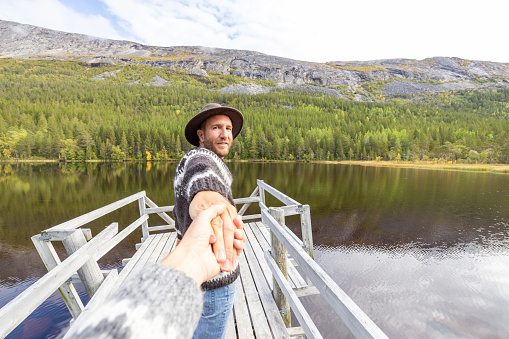 Man leading the way holding his partner's hand to a wooden pier above lake in autumn
