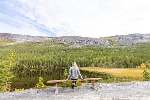Woman enjoying peaceful environment in Norway. Lake and green mountains.
She relaxes on a wooden bench