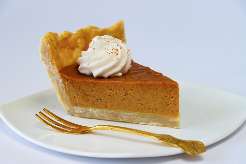 Stock photo showing close-up view of a slice of pumpkin tart topped with a swirl of whipped cream on a plate with a gold, dessert fork. Home baking concept.