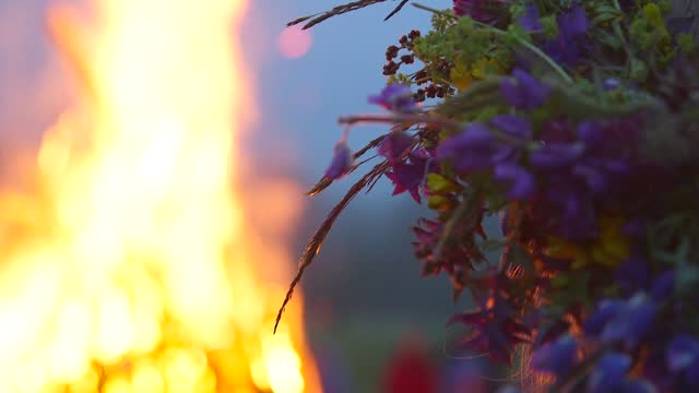 woman with a wreath of meadow flowers on her head watches a huge bonfire flame