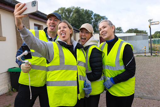 Three-quarter-length shot of a group of colleagues taking a silly selfie together, volunteering at a dog shelter located in Newcastle Upon Tyne. They are all wearing high-visibility jackets smiling at the phone.