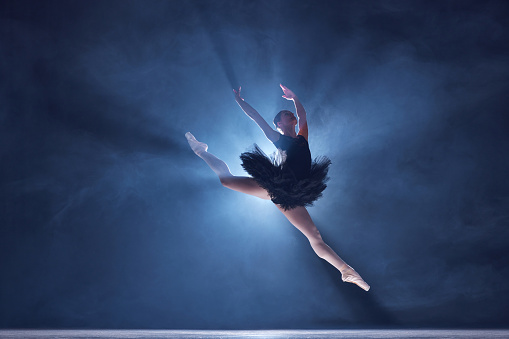 Graceful, attractive young woman, professional ballerina making performance, dancing on stage ever blue background with smoke effect. Concept of classical dance, art and grace, beauty, choreography