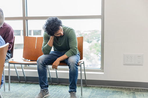 The exhausted young adult man sits in the waiting room of the community center to see a volunteer doctor during the health event.