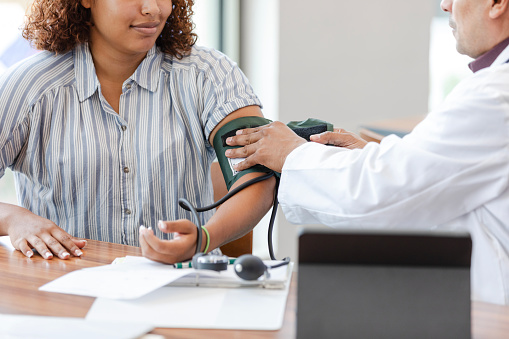 An unrecognizable male doctor wraps the blood pressure cuff around the unrecognizable young adult woman's arm.