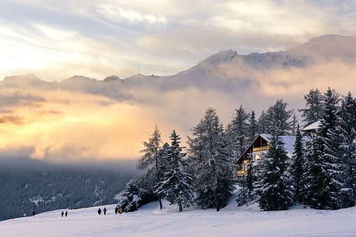 Winter landscape view of a mountain range partly covered by clouds and a chalet surrounded by trees at sunset, with hikers wandering in the snow