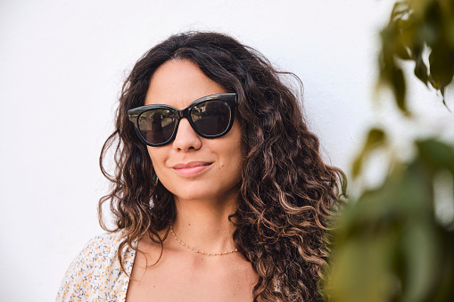 Retrato de chica joven con pelo negro y ondulado posando con gafas de sol frente a una pared blanca. Hojas de árbol a un lado.