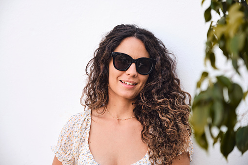 Retrato de chica joven con pelo negro y ondulado posando con gafas de sol frente a una pared blanca. Hojas de árbol a un lado.