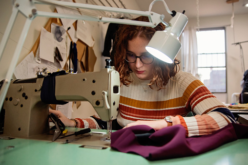 Young woman seamstress working in designer atelier at a sewing machine. She is in her twenties and is wearing a striped sweater and eyeglasses. Her hair is red and mid long. Horizontal waist up indoors shot with copy space. This was taken in Montreal, Quebec, Canada.